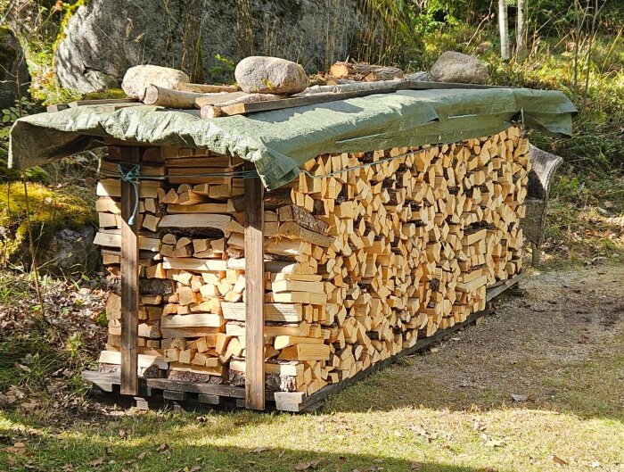 Travade vedklabbar täckta med grön presenning utomhus, cirka tre kubikmeter staplade.