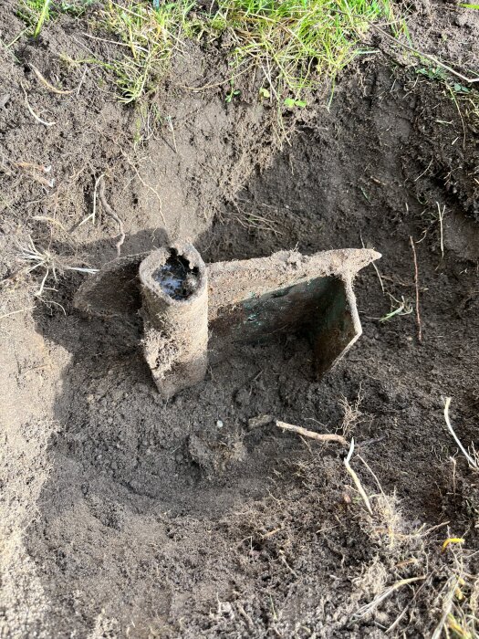 Nedgrävd metallbit och rör syns i en grop i en gräsmatta, omgiven av jord. Rörstycket ser rostigt ut mot den omgivande marken.
