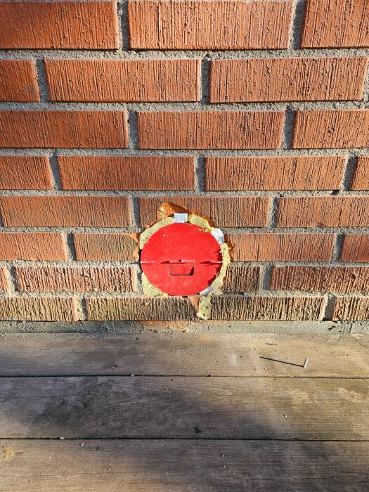 Röd ventilationsöppning på en tegelvägg, delvis täckt med isolering, sökes större kallrasskydd för att passa över öppningen.