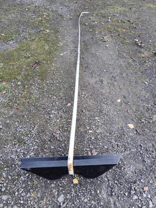 Självgjord moss-skrapa med ett blad fastsatt på en lång stång, liggande på grusig mark.