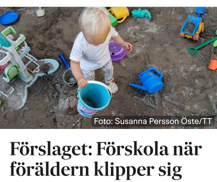 Ett barn leker i en sandlåda med spade, hink och andra leksaker, omgiven av sand och leksaksredskap.