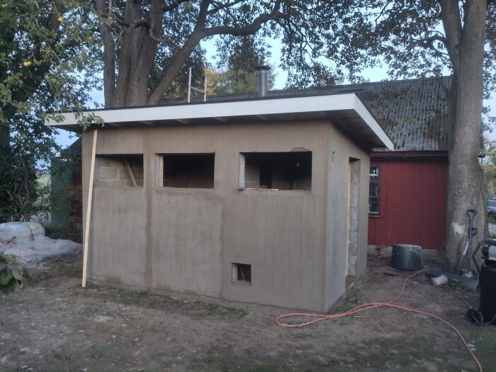 Litet nybyggt hus under träd, med putsade väggar, tre fönsteröppningar och en platt tak. Ett rött hus syns i bakgrunden.