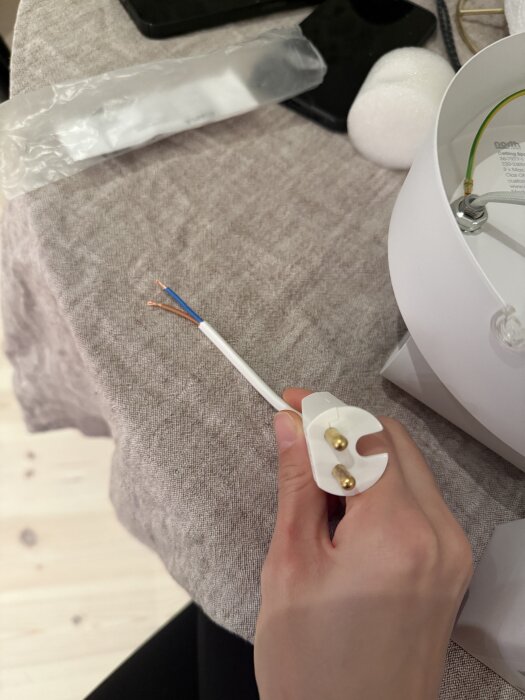 Hand som håller en elanslutning med blottade kablar och taklampa på ett bord täckt med tyg.