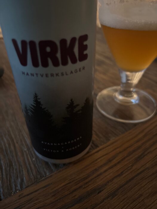 En burk Virke hantverkslager från Kvarnagården står bredvid ett glas gyllene öl med skumkrona på ett träbord.