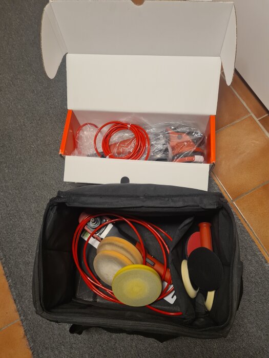 Röd poleringsmaskin med olika polerrondeller i en svart väska, bredvid en kartong med kablar och tillbehör. Perfekt för bilvård.