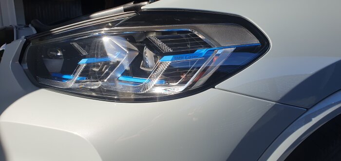 Närbild på en BMW-strålkastare med märkningen "BMW Laser", reflekterar ljus, monterad på en bil i grå färg.