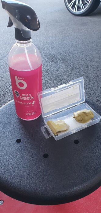 Sprayflaska med BBV Ultra Glide smörjmedel och en behållare med guldfärgat lermedel på en svart yta.