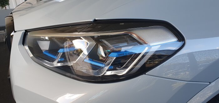 Närbild på en BMW strålkastare med laserinskription och detaljrik interiör, speglande lack av hög kvalitet på vit bil.