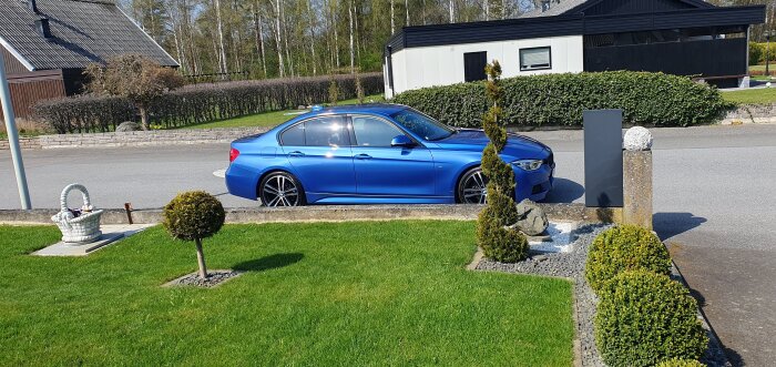 Blå bil parkerad på gata framför hus och häck, omgiven av gräsmatta och stensatta planteringar i trädgård. Solsken reflekteras på karossen.