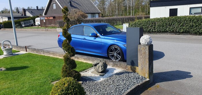 Blå bil parkerad vid en asfalterad gata framför ett hus, med välskött trädgård och dekorativa stenarrangemang i förgrunden.