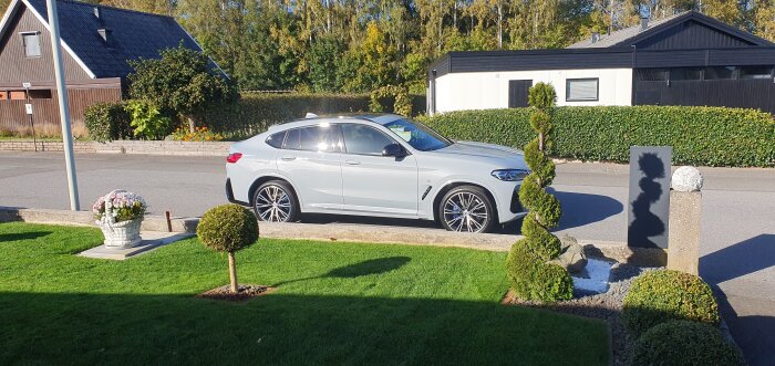 Vit bil parkerad på gata framför hus i lugnt bostadsområde, omgiven av välskött trädgård och grönska.