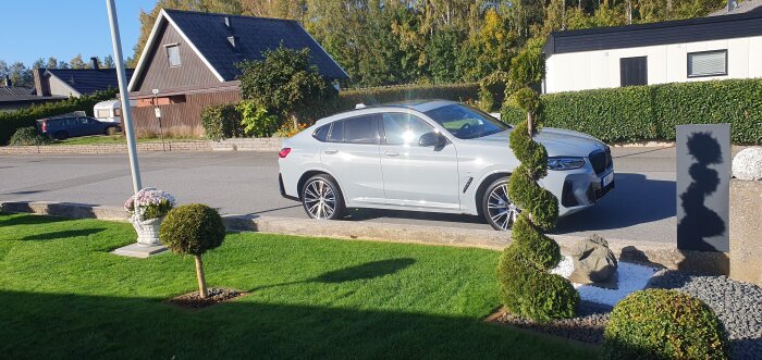 Vit bil parkerad vid väg, omgiven av välskött trädgård med buskar och blommor, soligt väder och villor i bakgrunden.