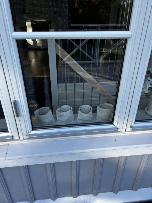 Fönsterram med flagnande färg på en byggnad, reflektion av man i rutig skjorta synlig i glaset, vita textilklädda ljuslyktor inuti.