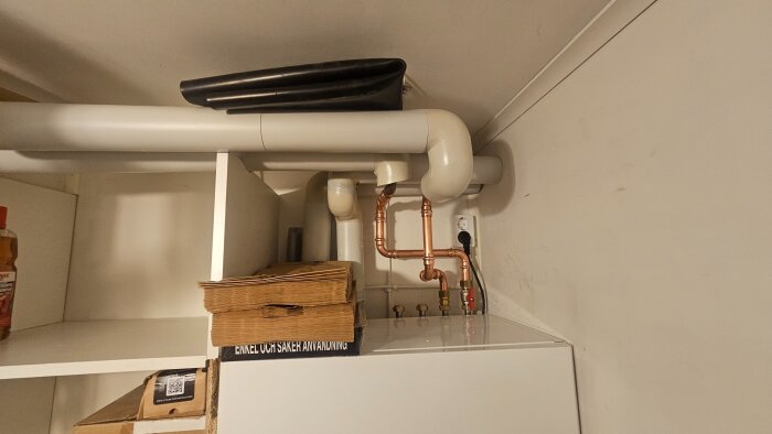 Rörinstallation i ett hörn av ett rum med kopparrör, ventil och hyllor med kartonger och en flaska städmedel.