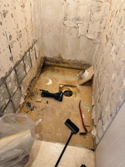 Rivet badrum med synliga fuktmärken, borttagen golvmatta, exponerad golvbrunn och verktyg; renovering påbörjad.