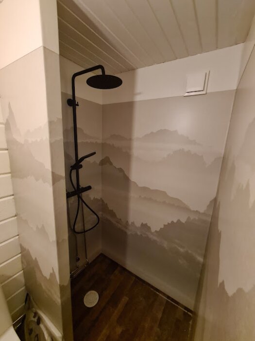 Duschhörna i ett badrum med våtrumsmatta och mönster av bergslandskap på väggarna. Mörk duscharmatur och trägolv.