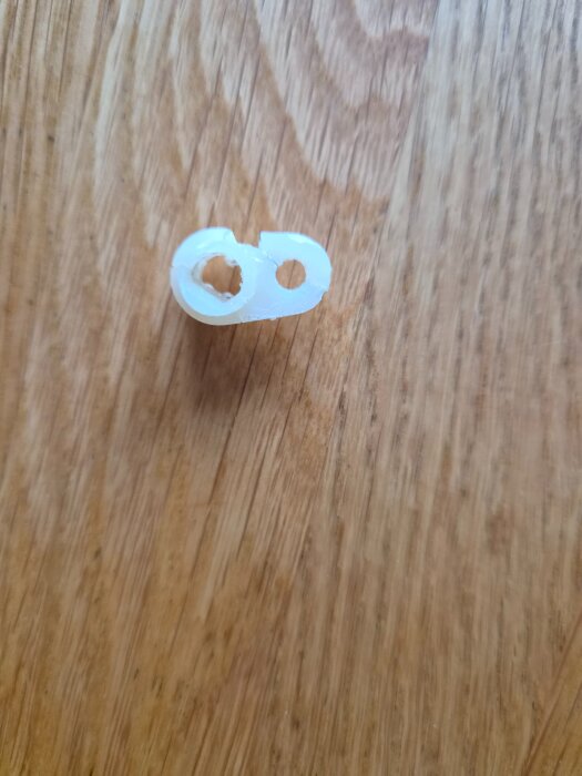 Trasig vit plastbit med två hål, avsedd att sitta mellan fönsterbroms och spanjolett på en Assa-fönsterbeslag.