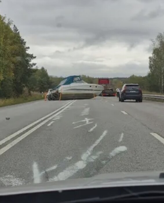 Båt blockerar väg efter att ha ramlat av släpvagn, med bilar stoppade bakom på landsväg.