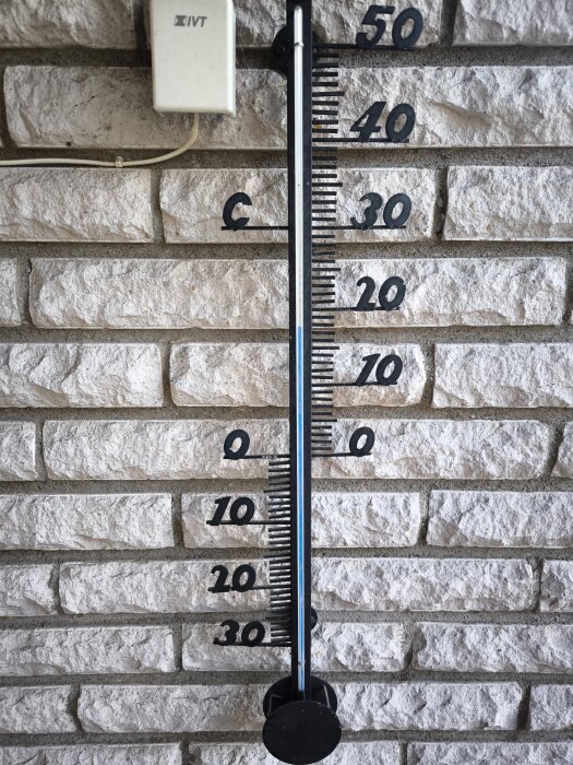 Utomhustermometer monterad på en vit tegelvägg, visar temperatur från -30 till +50 grader Celsius.