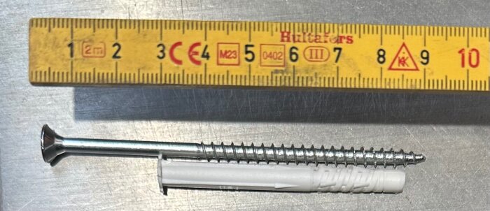 Skruv och plugg jämte en hopfällbar meterstock på metallbord. Skruven är silverfärgad och pluggen vit. Meterstocken visar skalan 1-10 cm.
