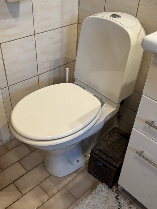 Gustavsberg toalett med vit sits och spolknapp, omgiven av beige kakel och en brun korg på badrumsgolvet.