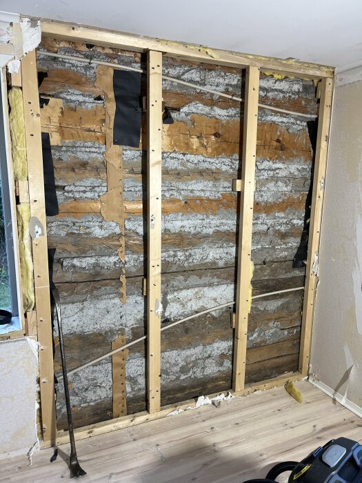 Renovering av timmervägg, visande blottade stockar, isoleringsmaterial och träreglar i ett rum efter borttagning av gammal väggbeklädnad.