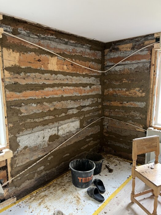 Renovering av hörnvägg med synliga timmerstockar, lerklining, och elkablar; golvet täckt av skyddspapper med hinkar och tofflor i förgrunden.