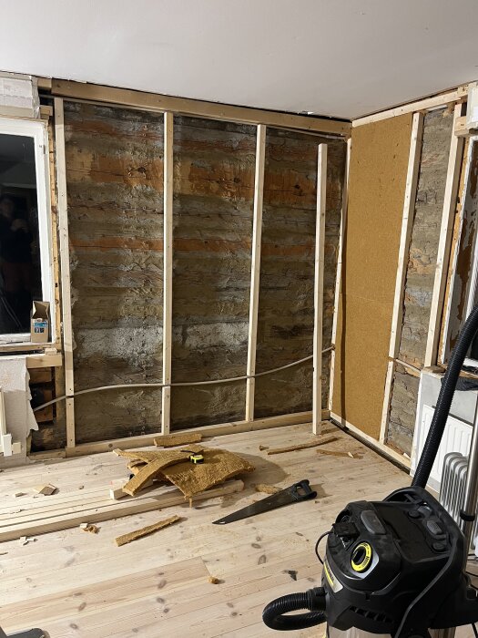 Renovering av vägg med synliga träreglar och timmervägg, verktyg och material på golvet, vakuum står i förgrunden.