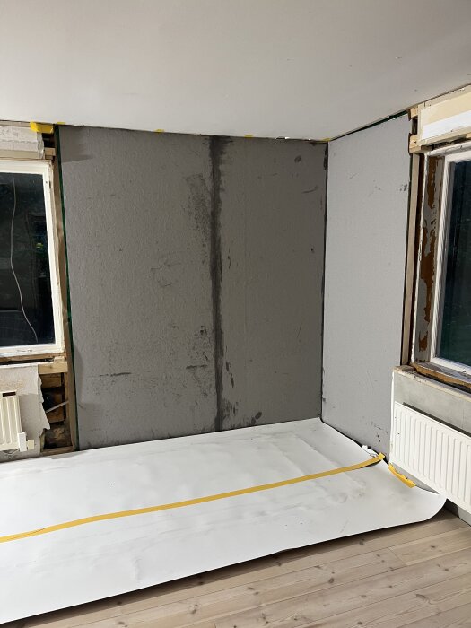 Vägg hörn under renovering med ny målning och underlagspapp på golvet. Fönster och element synliga, med öppnat lager tapet vid fönstret.