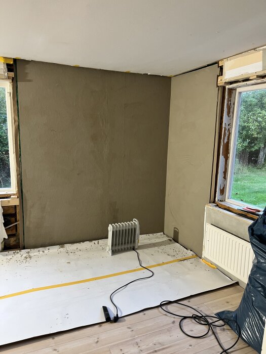 En målad vägg i ett rum under renovering, med en elradiator på golvet och delar av rummet fortfarande oisolerade.