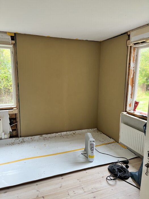 Renoverat hörn i rum med nymålade väggar i beige, omålade fönsterkarmar och golvskyddspapp. Element och elkabel syns på golvet.