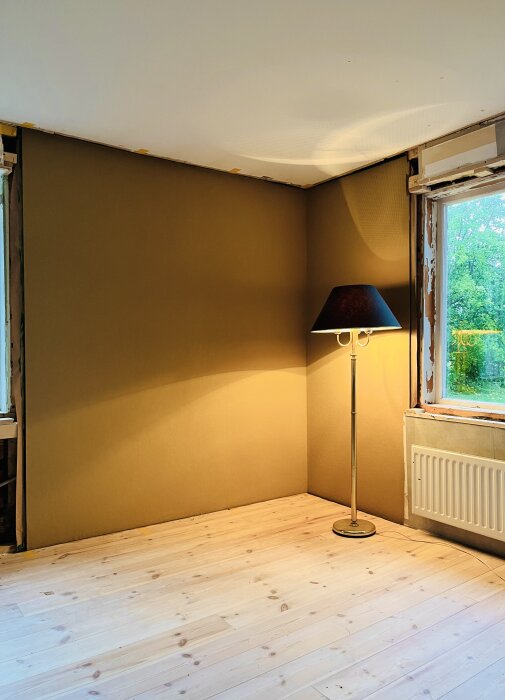 Renoverat hörn med nymålad vägg i en brun nyans, trägolv, golvlampa och del av fönster med träram under renovering.