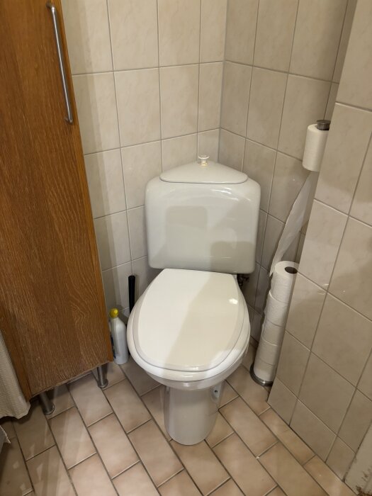 Sliten gammal toalett i ett hörn med vitt kakel och beige golvplattor; ett skåp och toalettpapper syns bredvid.