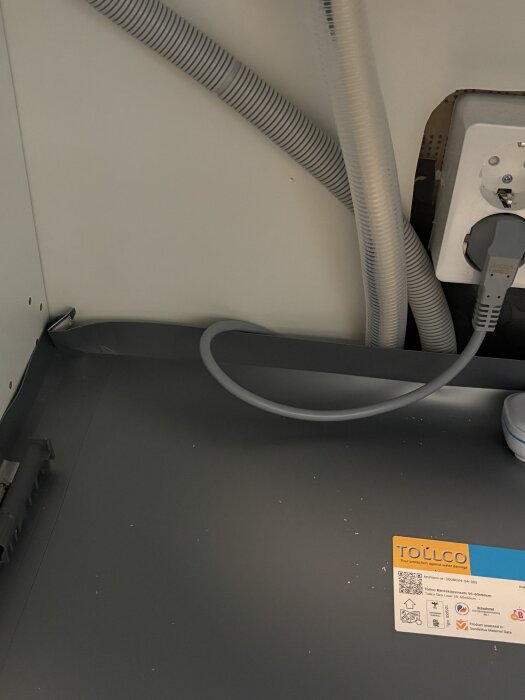 Diskho under skåp med vattenledningar och eluttag, synlig tätning runt avlopp och plastmatta som inte når hela vägen bak.