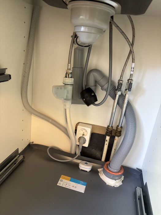 Vatteninstallation under diskho med synliga rör och kablar, slarvigt tätade avloppsanslutningar och plastmatta som inte når väggen.