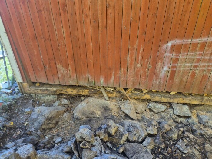 Röd träpanelvägg med skador längs basen, omgiven av stenar och jord.