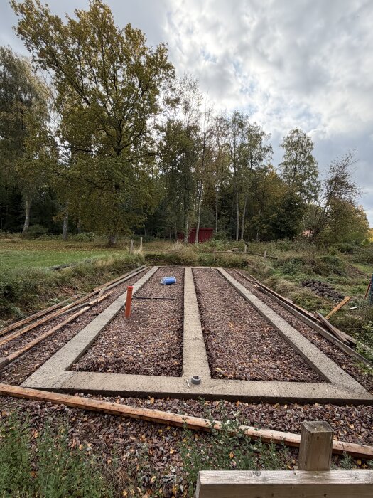 Platsgjuten betongsula i skogsmiljö med makadam mellan sektionerna. En blå plastbehållare och röd rördel syns på sulan.