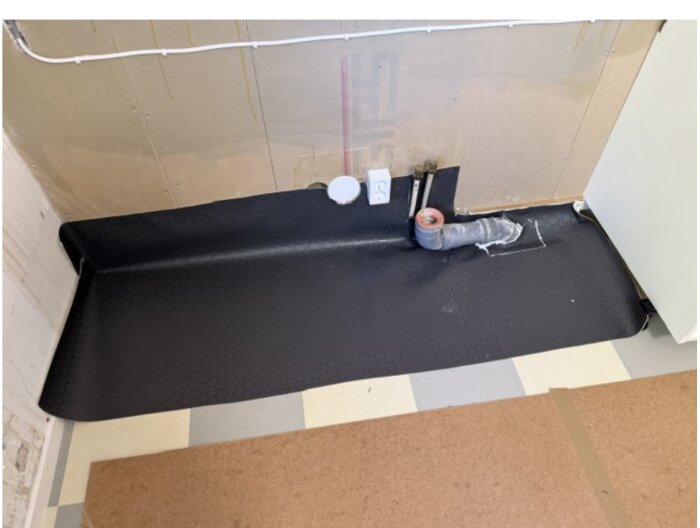 Svart matta med uppvik vid vägg, rörgenomföring och kopplingar i rum under renovering.