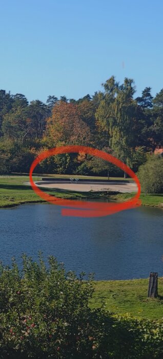 Sjön med grönska och en betongstruktur i bakgrunden, markerad med en röd cirkel.