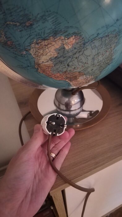 Hand som håller en kontakt bredvid en jordglob på ett bord, globen har bronsfärgad bas och visar del av jorden där Nord- och Sydamerika syns.