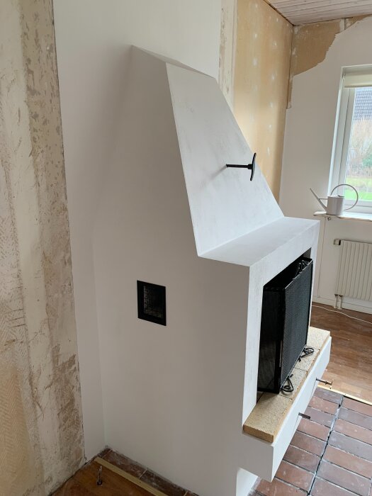 Putsad vit kamin i ett rum under renovering, med en svart ventil och eldfast glasyta, omgiven av exponerade väggar och ett fönster med utsikt.
