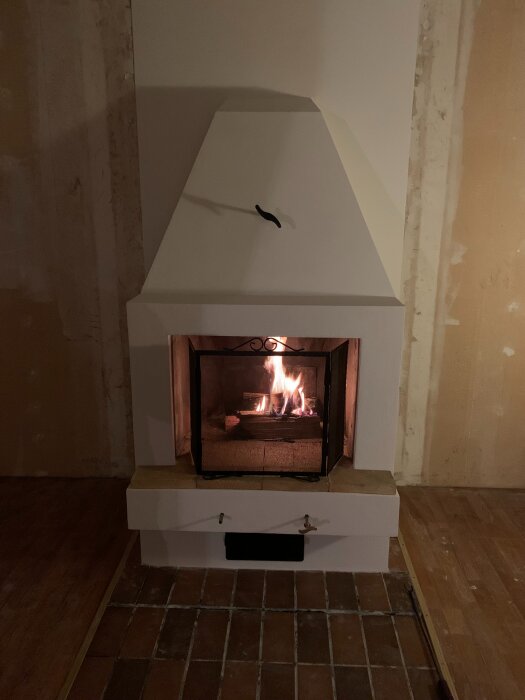 Öppen spis med eld brinnande inuti, omgiven av vitputsade väggar och en tegelgolv i ett rum under renovering.
