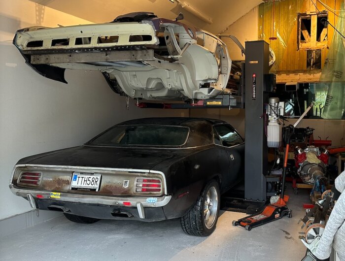 Bilprojekt på en flyttbar enpelarlyft i ett garage, med en gammal bil ovanpå och en annan under, omgiven av verkstadsutrustning.