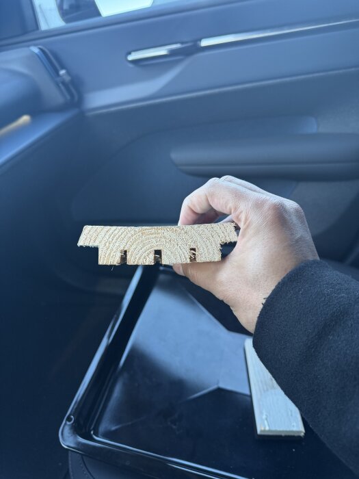 Hand håller en träbit med spår på kortsidan, mot en mörk bakgrund i en bil.