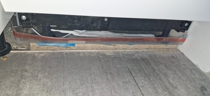 Installationen av uppsamlingstråg under diskmaskinen, med synliga skruvar och ett vitt vattenlarm på plats.