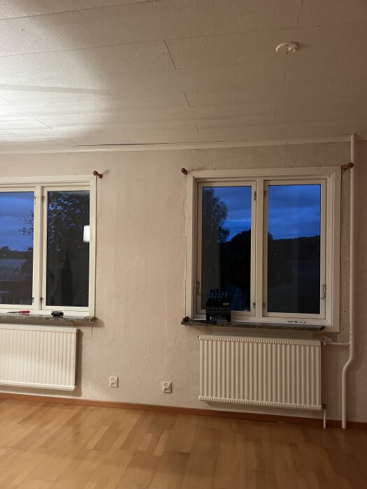 Rum med två stora fönster, beige tapet och trägolv. Tapeten är delvis borttagen, synlig underlag på väggen. Två element under fönstren.