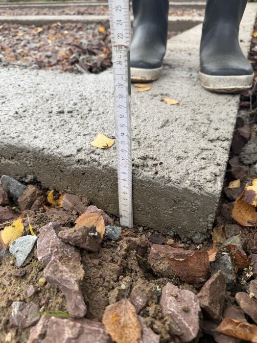 Mätning av tjockleken på en betongsula, omgiven av jord och grus, med ett måttband; stövlar syns i bakgrunden.