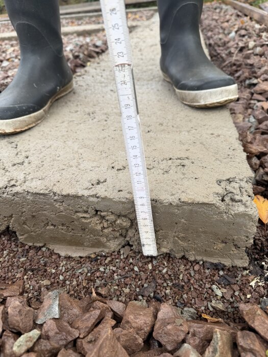 Närbild av en betongsula med måttstock och grus runtomkring, visar sulans bredd och höjd. Person i stövlar står på sulan.