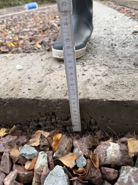 Närbild av betongsula mätt med måttstock vid kanten ovanpå ett lager grus och löv. En person i stövel står bredvid.