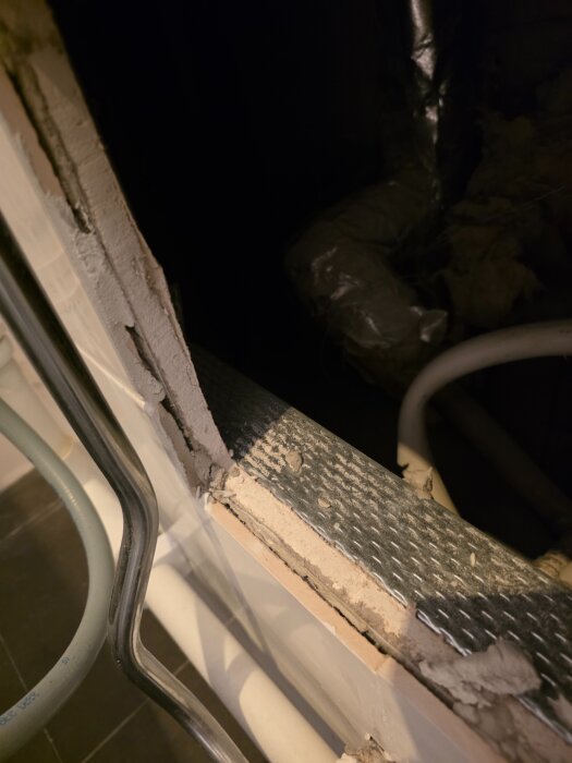 En halvfärdig väggöppning med synliga rör och hörn av byggmaterial täckt med metall och murbruk. Belysning från sidan lyser upp strukturen.